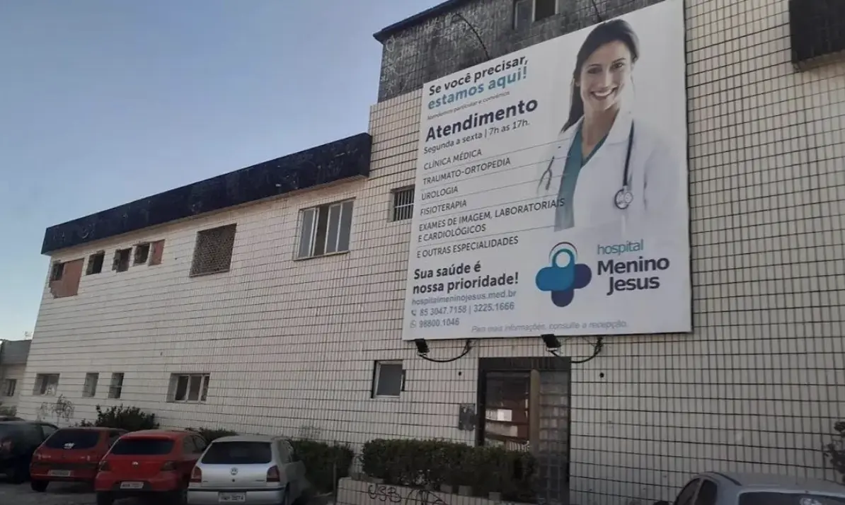 Fachada de um prédio ladrilhado bege com uma espécie de outdoor com informações sobre o atendimento, horário e modalidades do Hospital Menino Jesus, parte da rede referenciada Bradesco Saúde em Fortaleza