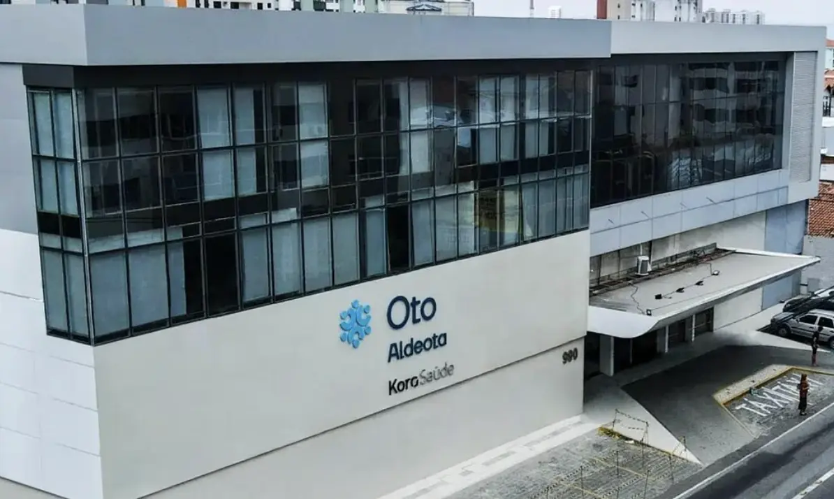 Foto da fachada de um hospital com as paredes todas envidraçadas no andar superior e paredes em cor cinza claro no térreo. Na fachada , é possível ler em letras grandes azuladas: "Oto Aldeota" KoraSaúde, se trata de um hospital referenciado do Bradesco Saúde em Fortaleza