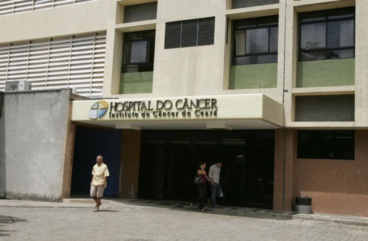 entrada de um prédio bege com janelas verdes, a foto mostra duas pessoas caminhando pela calçada e um homem atravessando a rua. A entrada tem uma fachada avançada com o letreiro em alto relevo em letras grandes "Hospital o Câncer - Instituto do Câncer do Ceará" pertencente à rede de hospitais referênciados do Bradesco Saúde em Fortaleza