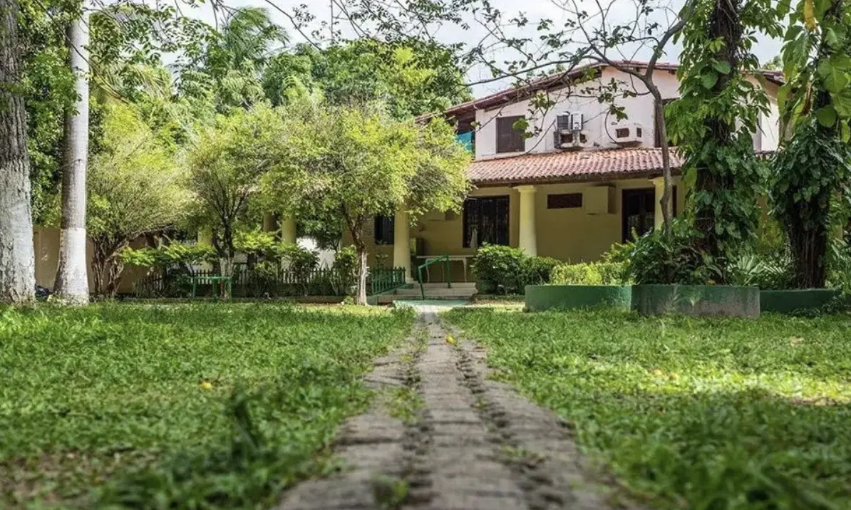 foto em alta resolução da entrada de uma clínia. A foto mostra uma trilha que vai até a porta da residência principal, uma residência grande de dois andares, a triha percorre um jardim com árvores e grama. Se trata da Clínica Vila Nova referenciada do Plano de Saúde Bradesco em Fortaleza